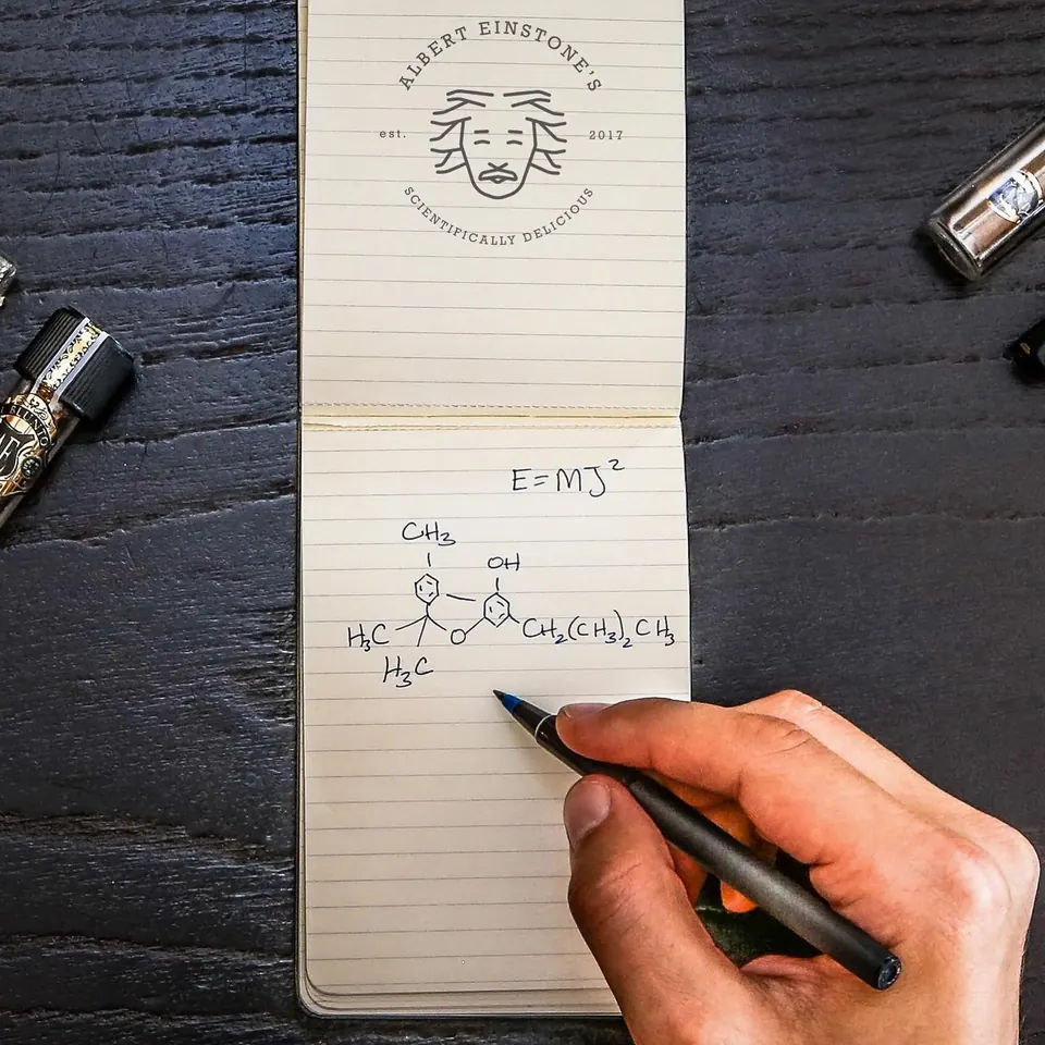 A hand writing E=MJ² and a cannabinoid structure in an Albert Einstone's notebook, surrounded by El Blunto tubes